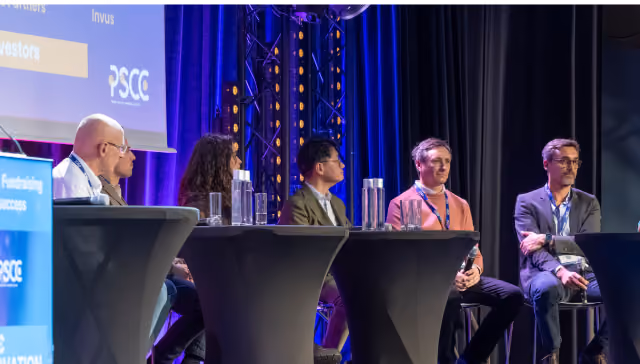 Five panelists seated behind high tables on stage during a conference discussion with blue and purple lighting.