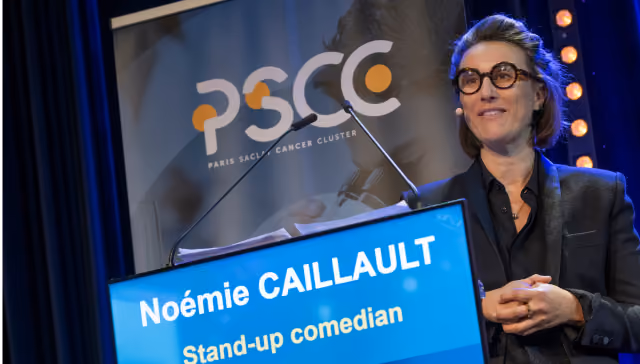 Woman with glasses speaking at a podium displaying name Noémie Caillault, stand-up comedian.