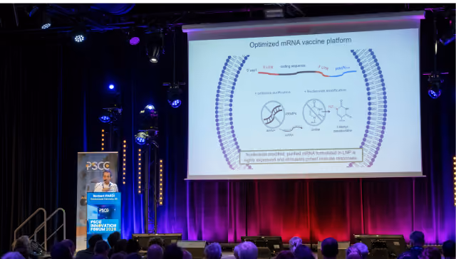 Speaker at a podium presenting a slide on optimized mRNA vaccine platform before an audience.