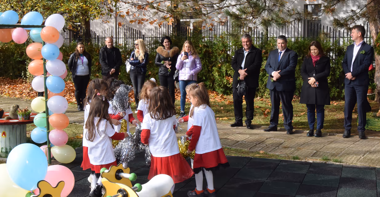 A group of young girls in coordinated white shirts and red skirts perform a dance or cheerleading routine in a playground decorated with balloons