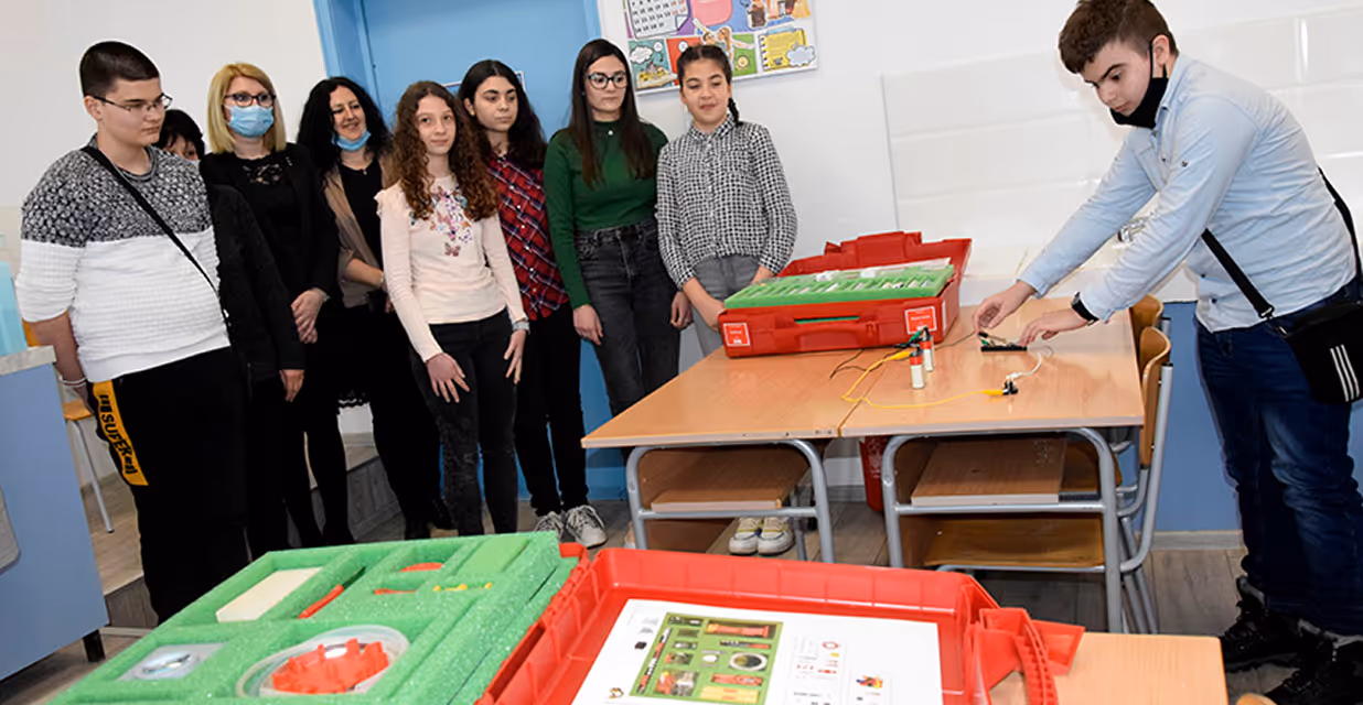 A boy performs an experiment with electrical wires at a classroom desk while other students and teachers observe, with science kits laid out on the desks