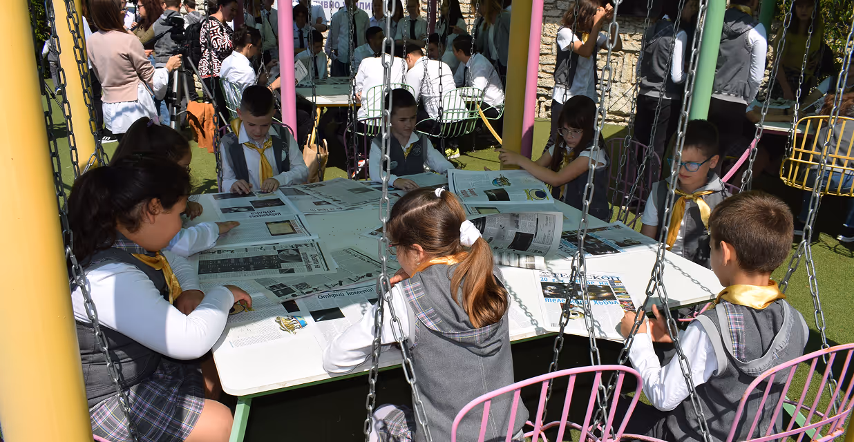 A group of preschool children stands with teachers outside near trees, with chess tables set up on benches and children’s artwork