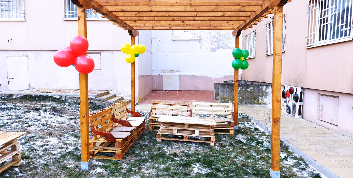 Outdoor wooden pergola with pallet furniture and festive balloons — A small courtyard features a newly constructed wooden pergola adorned with red, yellow, and green balloons