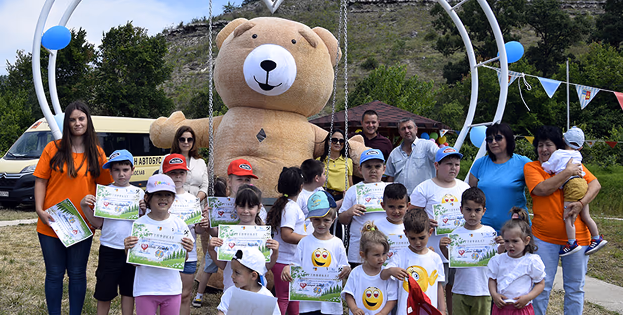Outdoor children's event with large teddy bear mascot and certificate ceremony – Group of children holding certificates, standing in front of a giant teddy bear figure under a decorative arch, accompanied by teachers and parents, celebrating a school or community achievement event in a grassy outdoor area.