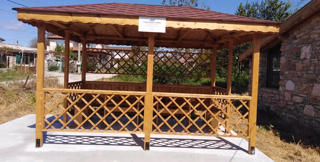 Wooden gazebo built for community use with Energo-Pro sign – A newly constructed wooden gazebo with lattice fencing and benches, placed on a concrete base in a rural village setting, marked with an “ENERGO-PRO” plaque, serving as a shaded communal gathering or learning space.