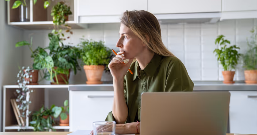 Thoughtful woman standing in front of laptop with pen and documents in her hands