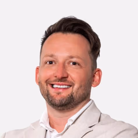 Lächelnder Mann mit kurzem braunem Haar, Bart und hellgrauem Sakko vor weißem Hintergrund.