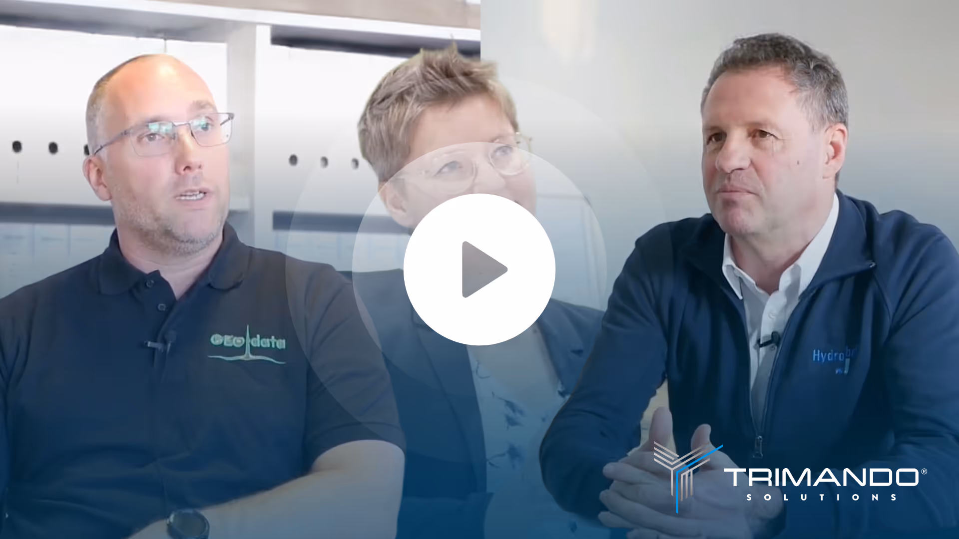 Drei Personen sitzen zu einem Interview in einem modernen Büro, zwei Männer und eine Frau in der Mitte, mit Firmenlogos auf ihren Kleidern.