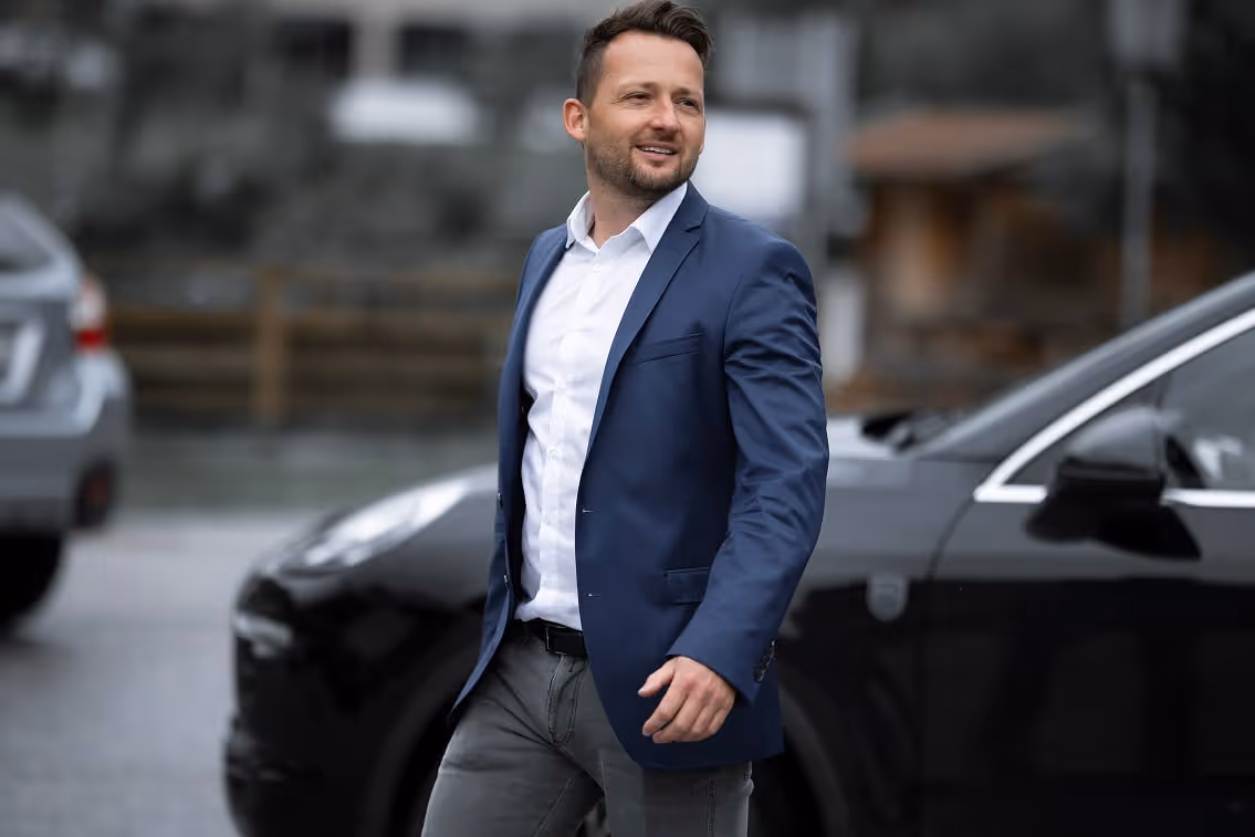 Ein lächelnder Mann mit kurzem Haar, der einen blauen Blazer und ein weißes Hemd trägt, geht an schwarzen Autos im Freien vorbei.