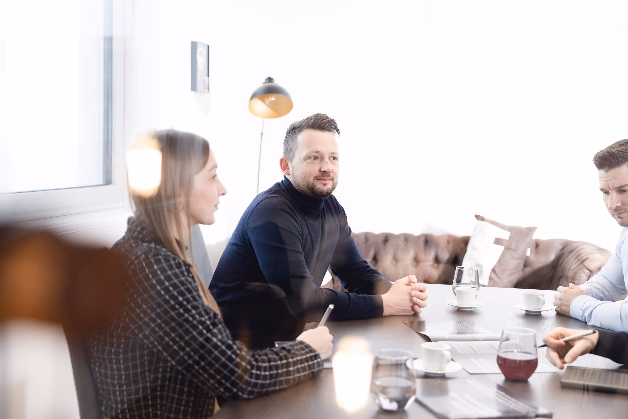 Drei Personen führen eine Besprechung an einem Tisch mit Kaffees und Dokumenten in einem hellen Büro.