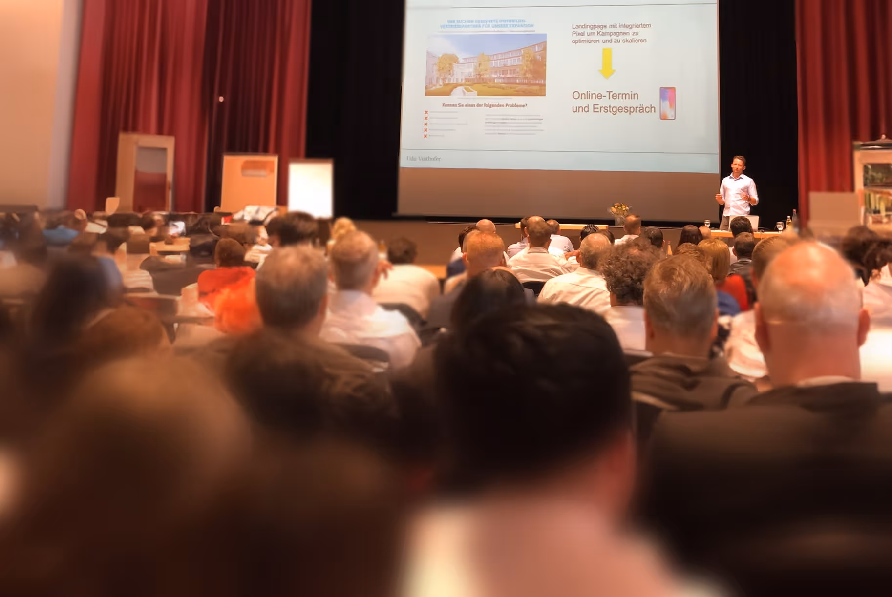 Ein Redner hält einen Vortrag vor Publikum in einem Veranstaltungsraum mit einer Präsentation auf einer großen Leinwand.