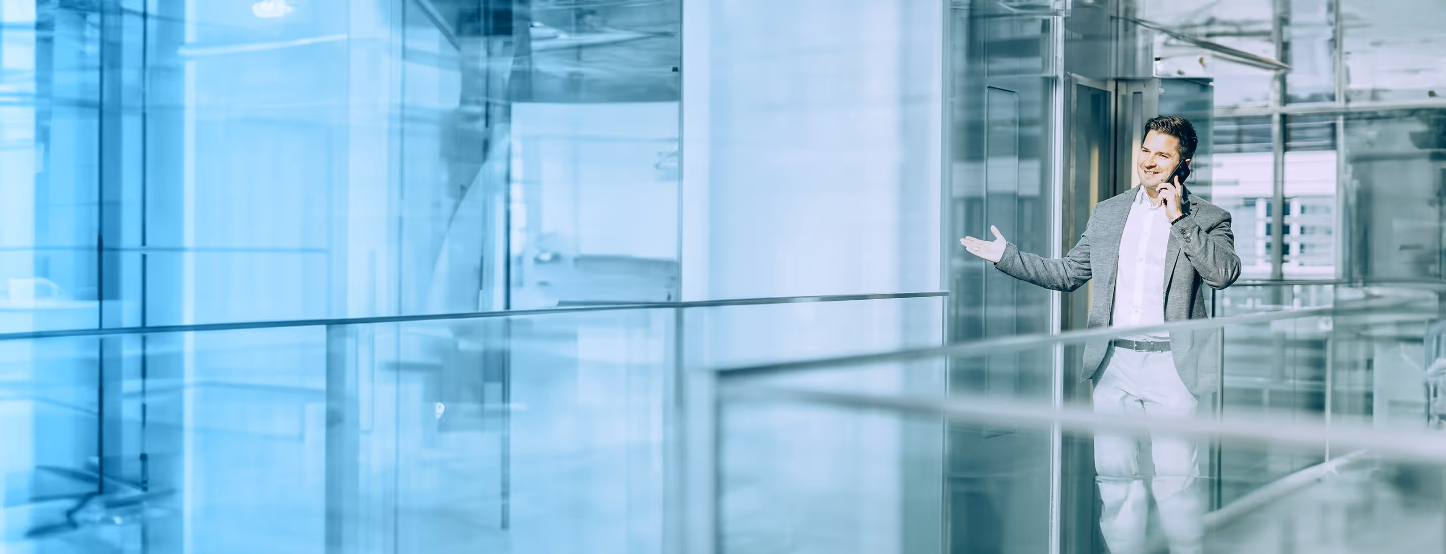 Geschäftsmann in grauem Sakko spricht lächelnd am Smartphone in einem modernen, gläsernen Bürogebäude.