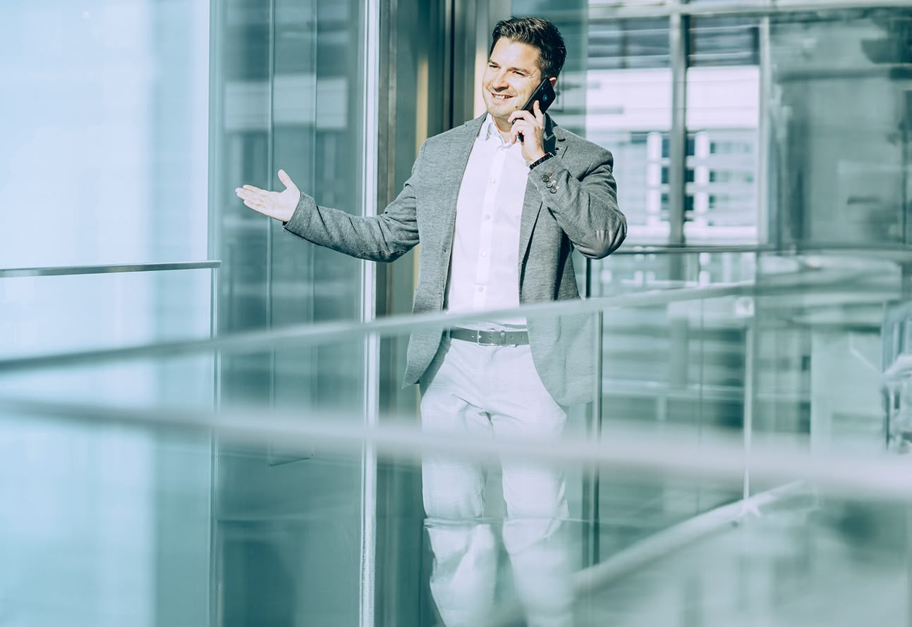 Geschäftsmann im grauen Sakko spricht telefonierend und gestikuliert in einem modernen Bürogebäude.