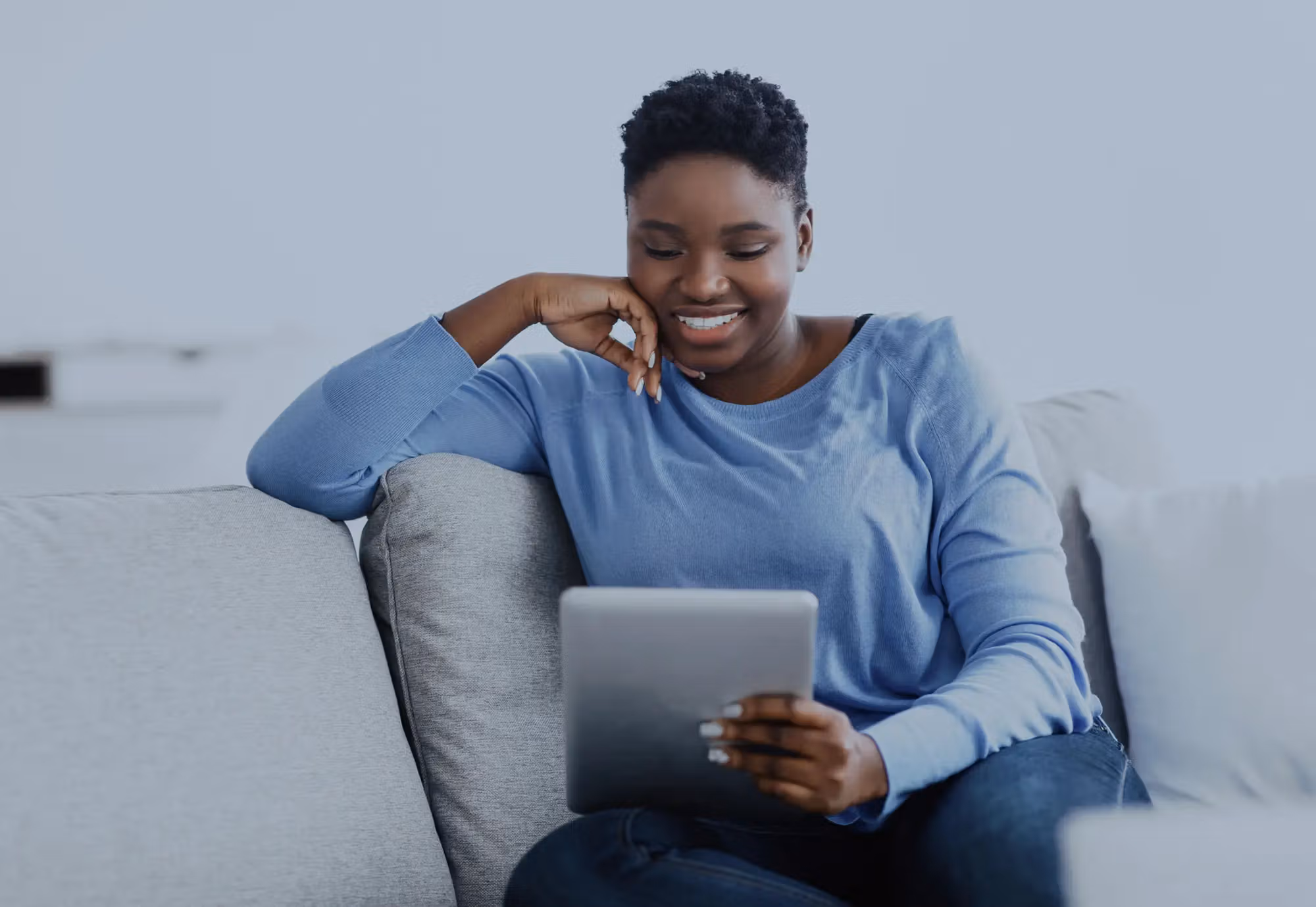 Woman in blue sweater smiling while using tablet on comfortable couch