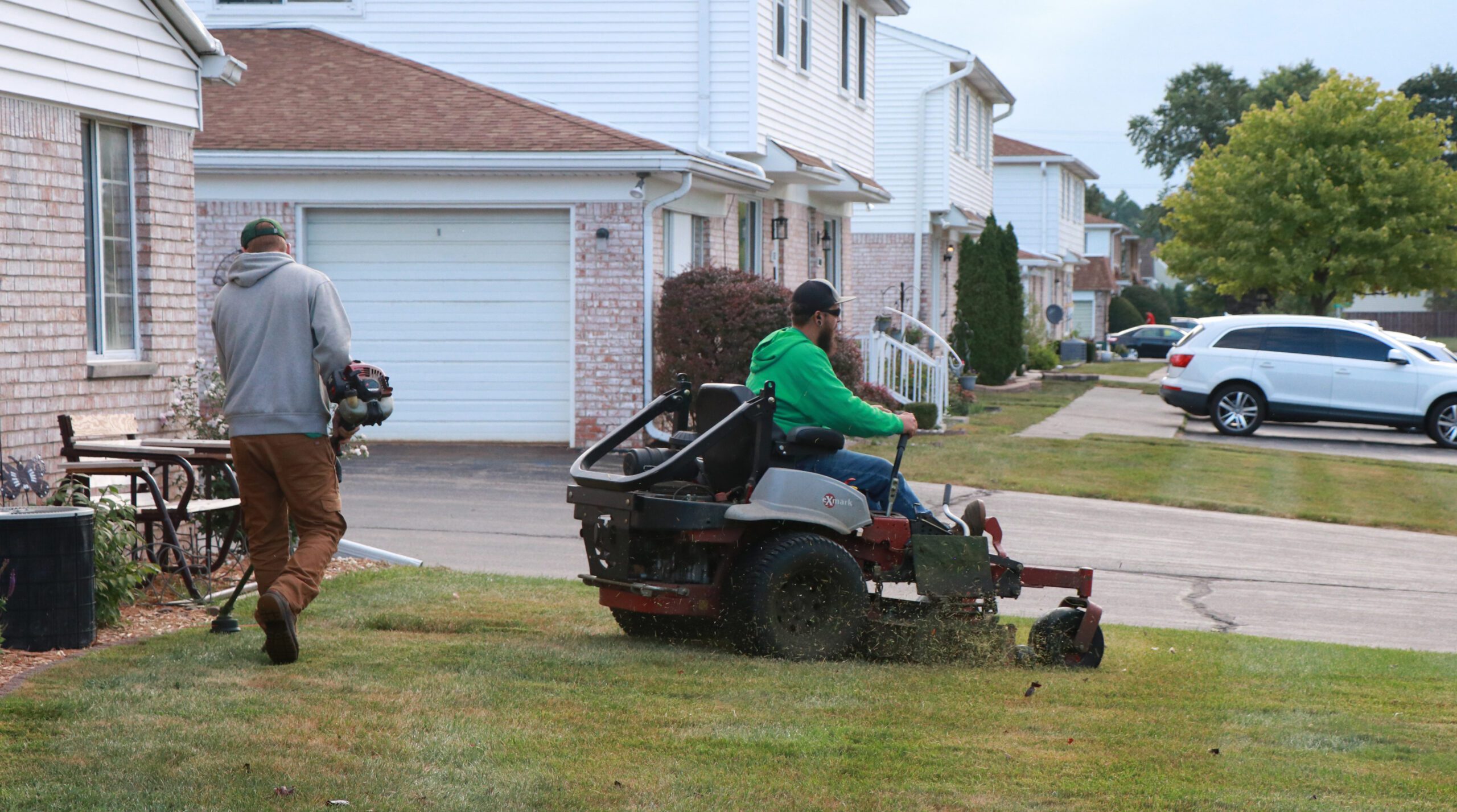 Two landscapers mowing suburban lawn with riding and handheld lawn mowers