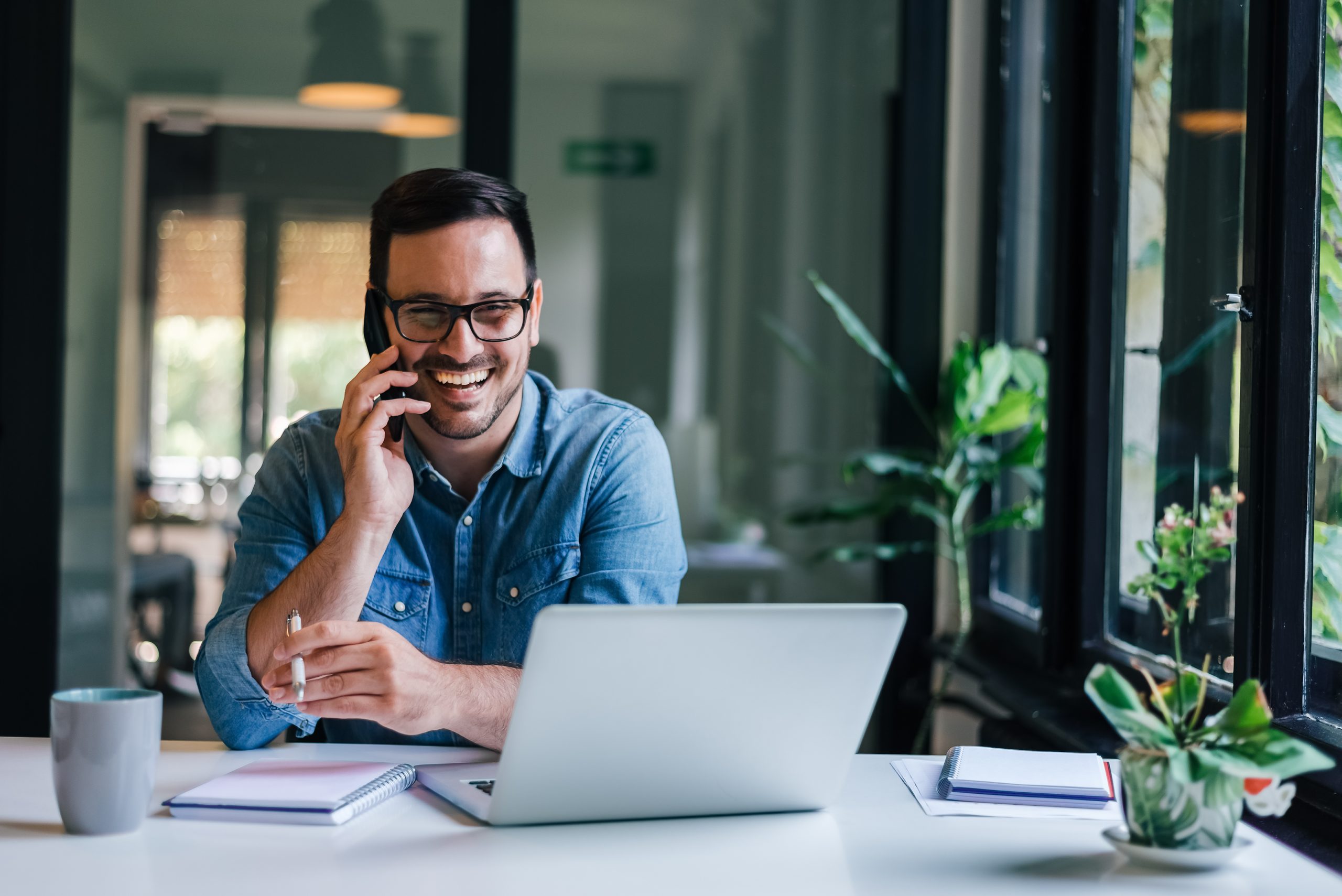 Smiling man in denim shirt working on laptop and talking on phone