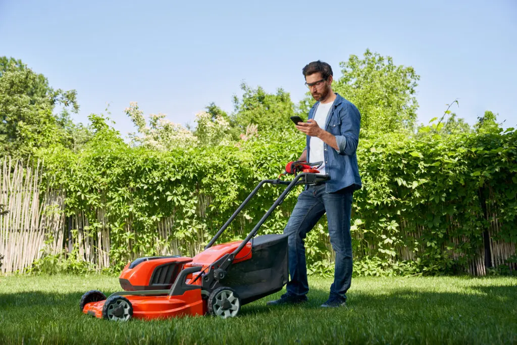 Man checks phone while using red electric lawn mower in backyard