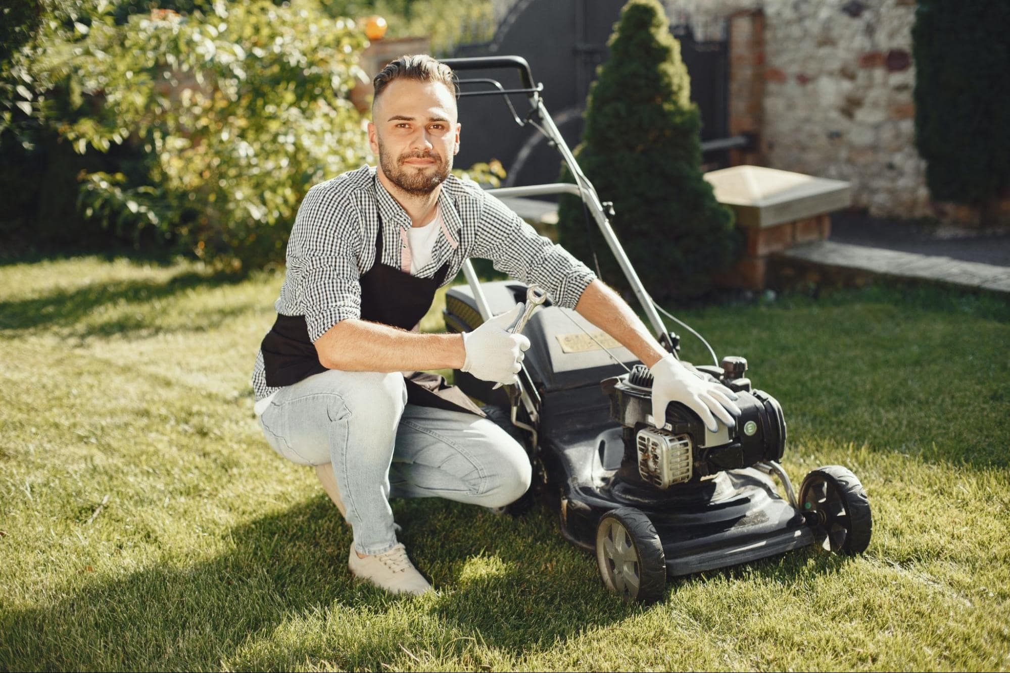 Person in checkered shirt kneeling next to lawn mower in backyard garden