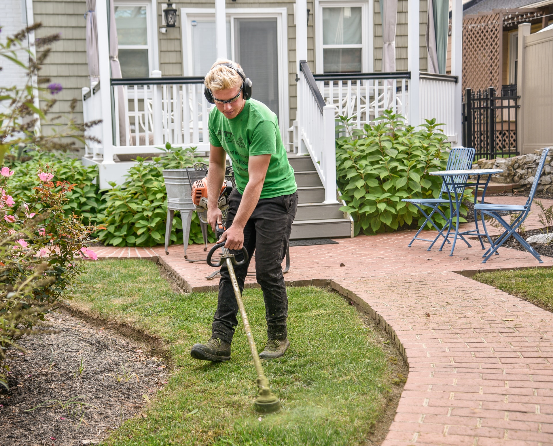Person in green shirt using string trimmer to edge lawn near brick path