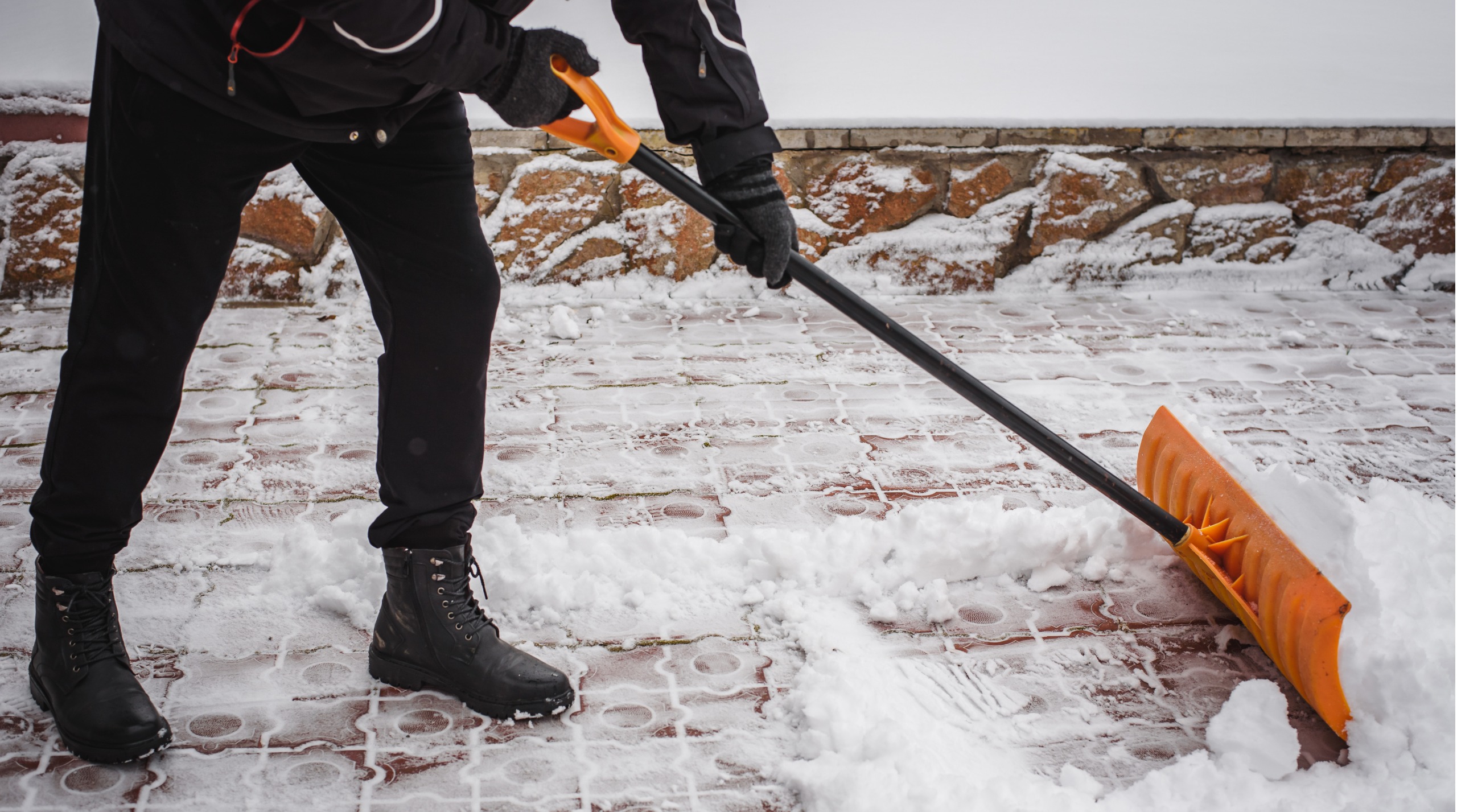 Person clearing snow with orange snow shovel on brick paved ground