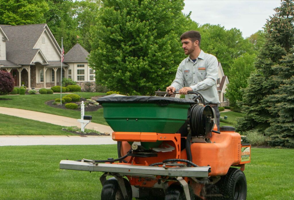 Landscaper operating orange and green ride-on spreader on suburban lawn