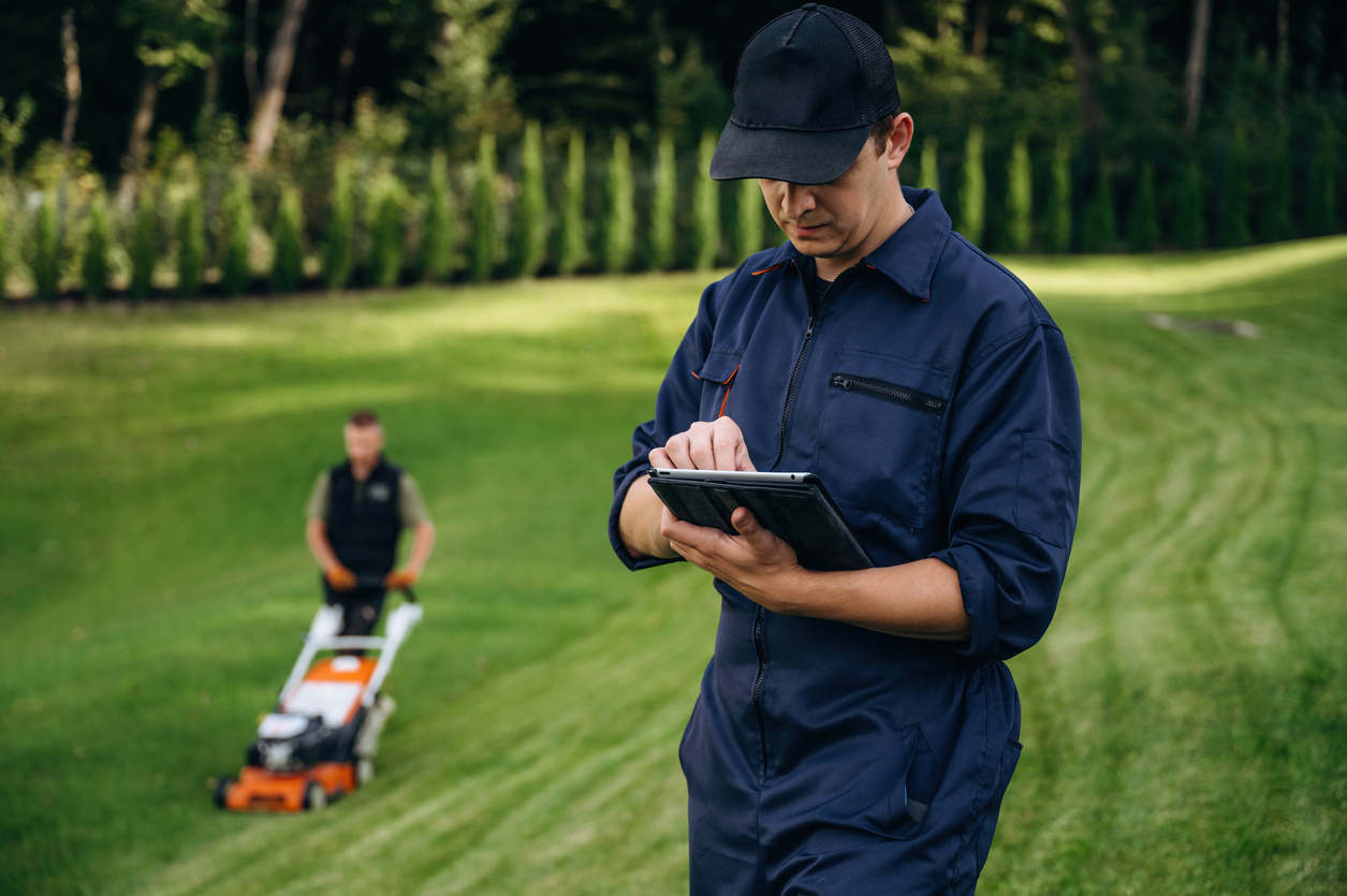 Landscaper using tablet while colleague mows lawn in background