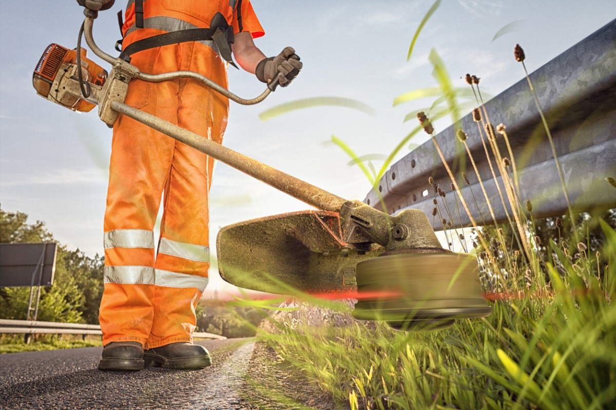 Worker in orange safety gear using string trimmer along roadside guardrail