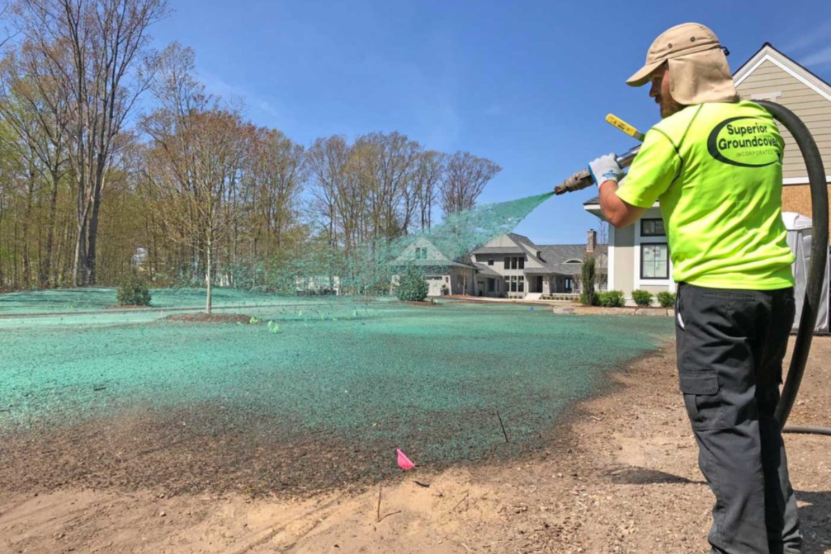 Worker spraying green hydroseeding mixture on bare soil near residential home