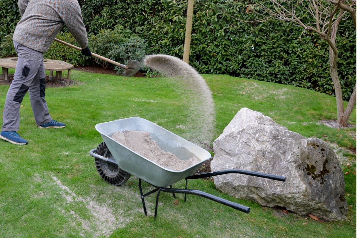 Person shoveling sand into wheelbarrow near large rock in garden