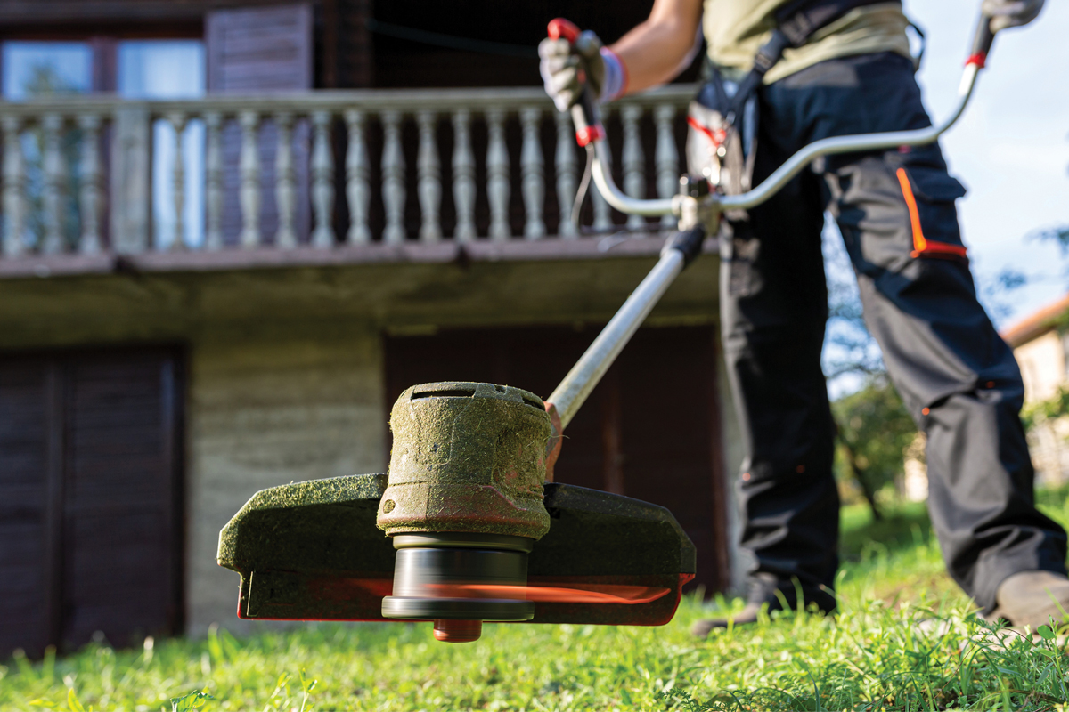 Person using string trimmer to cut grass near wooden house