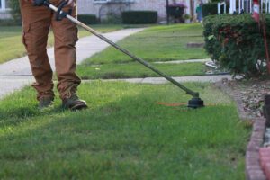 Person using string trimmer to edge grass along sidewalk and garden