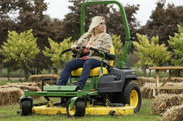 Woman riding green and yellow zero-turn lawn mower in orchard