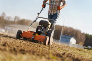 Person using a power rake to prepare soil in a grassy outdoor area