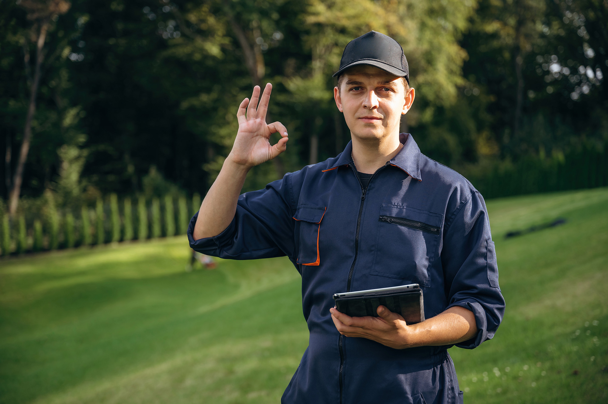 Worker in blue uniform makes okay sign, holding tablet outdoors