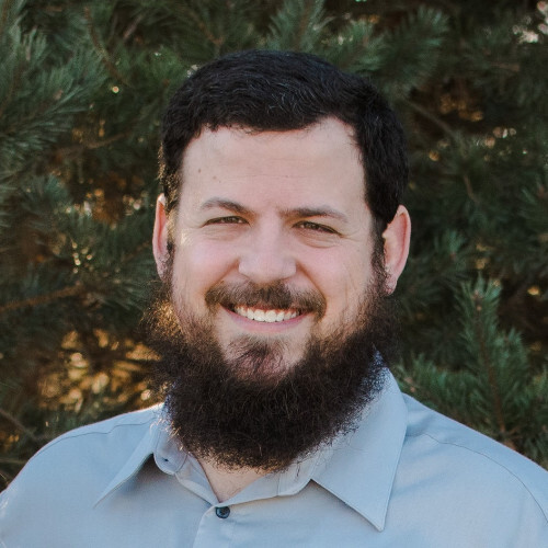 Smiling man with full beard standing in front of green pine trees