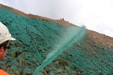 Worker sprays green hydroseeding solution on rocky hillside slope