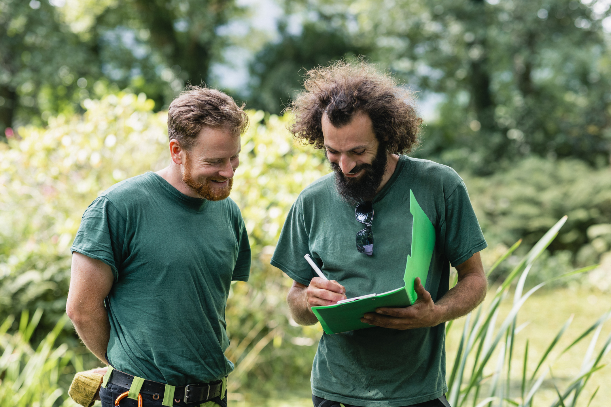 Two gardeners in green shirts collaborate outdoors, taking notes in nature