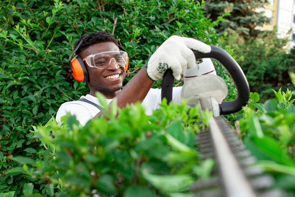 Smiling worker trimming hedges with protective gear in green garden