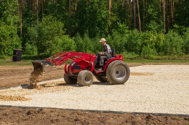Red tractor spreading gravel on construction site with forest background