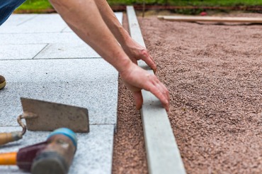 Hands laying paver stone on sandy ground with construction tools nearby