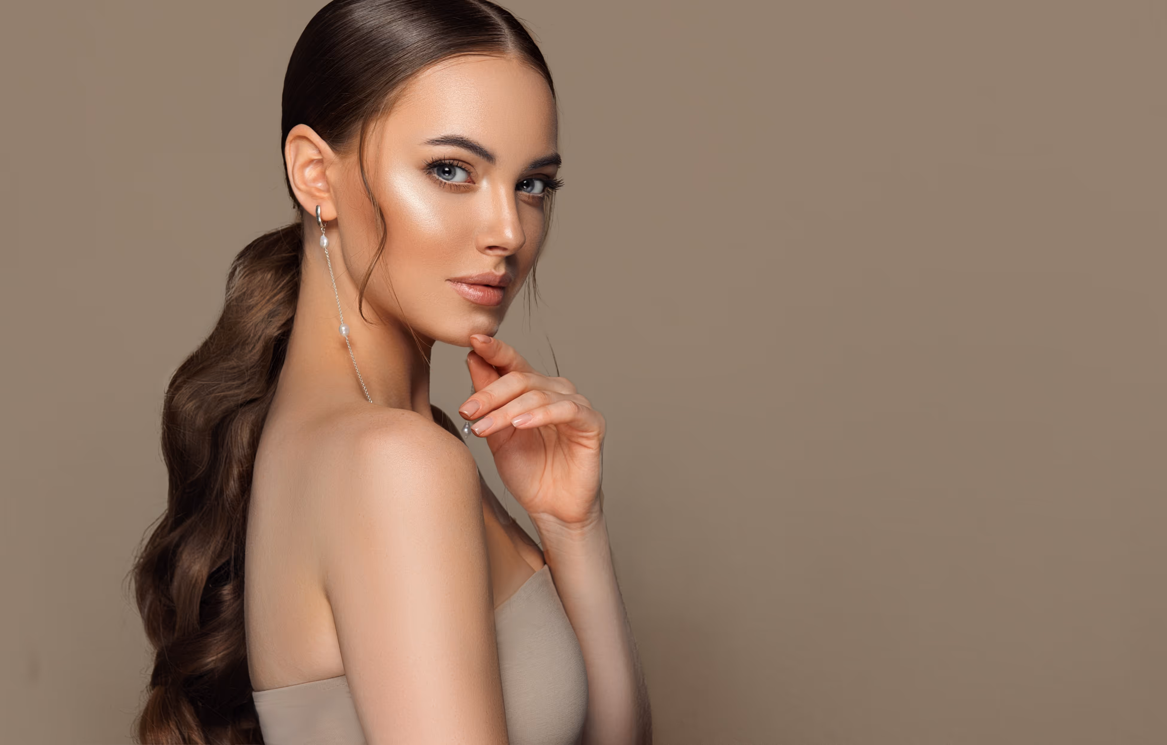 Portrait of a woman with long wavy brown hair tied back, wearing pearl earrings and a strapless beige top, posing against a plain beige background.