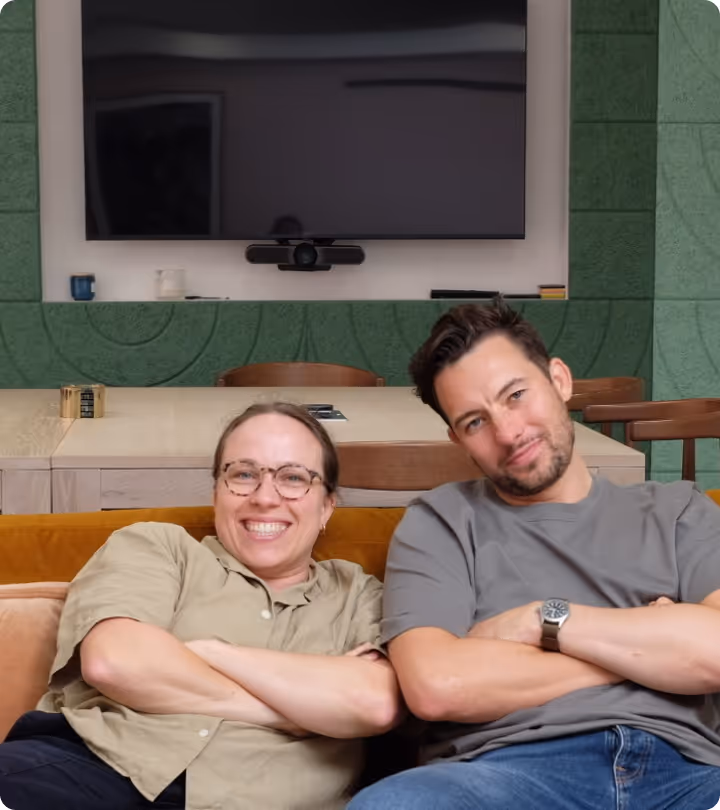 Smiling woman with glasses and man with beard sitting side by side on a yellow couch with arms crossed.