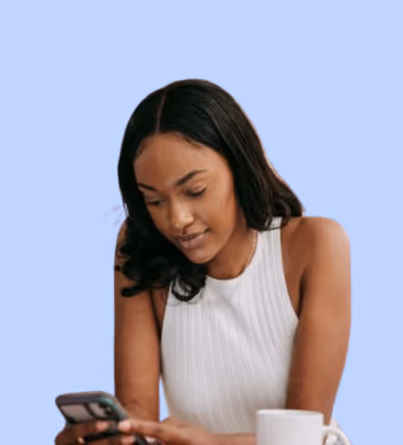 Woman with long dark hair wearing a white sleeveless top looking at her smartphone with a white coffee mug in front of her.