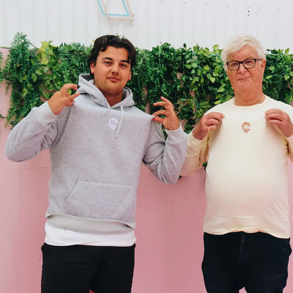 Two men showing off Oma's Soep branded merchandise, one wearing a grey hoodie and the other a cream longsleeve, posing in front of a pink wall with green plants