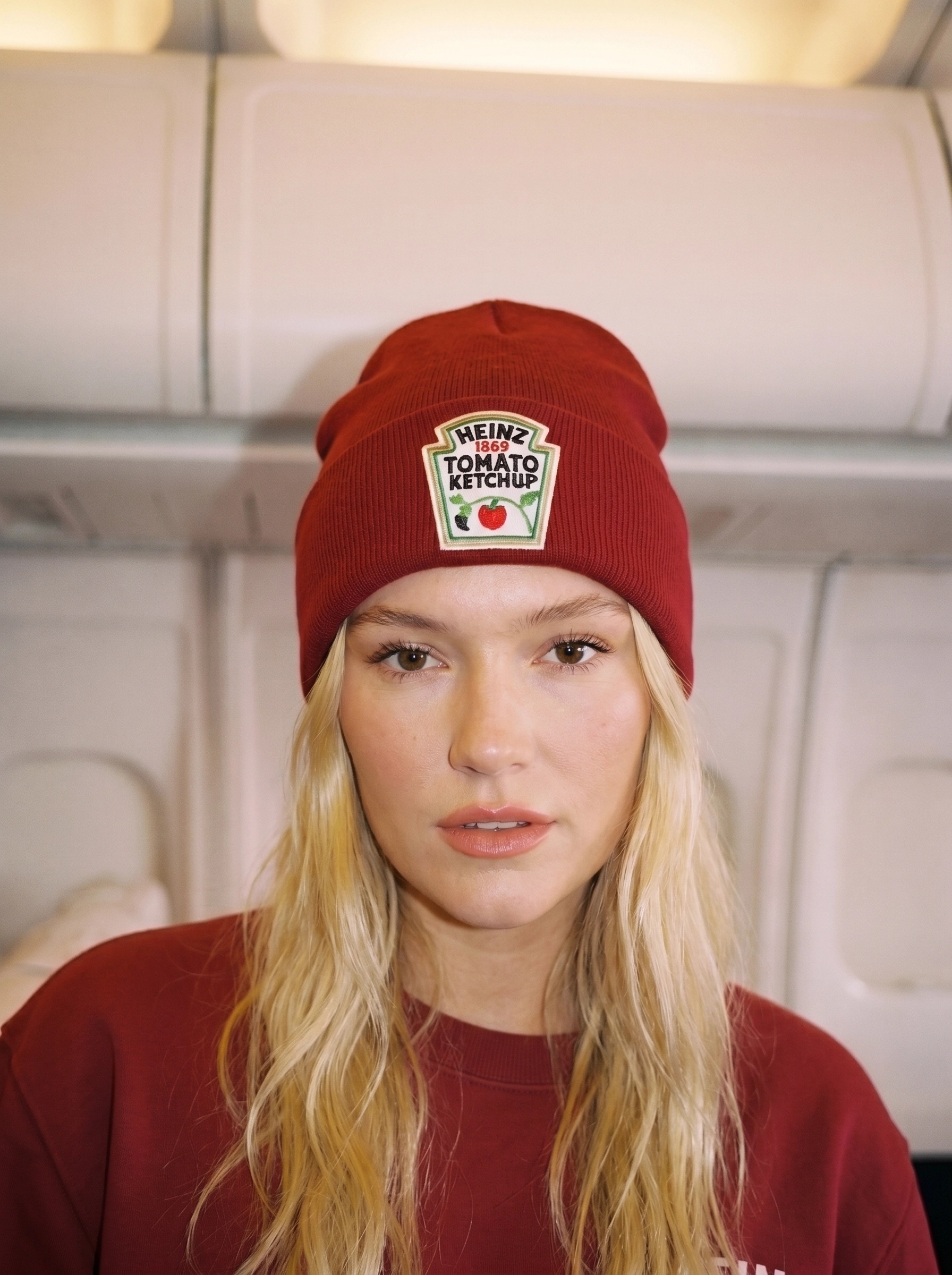 Close-up van model met bordeauxrode Heinz Tomato Ketchup beanie met geborduurde label-patch, in vliegtuigcabine