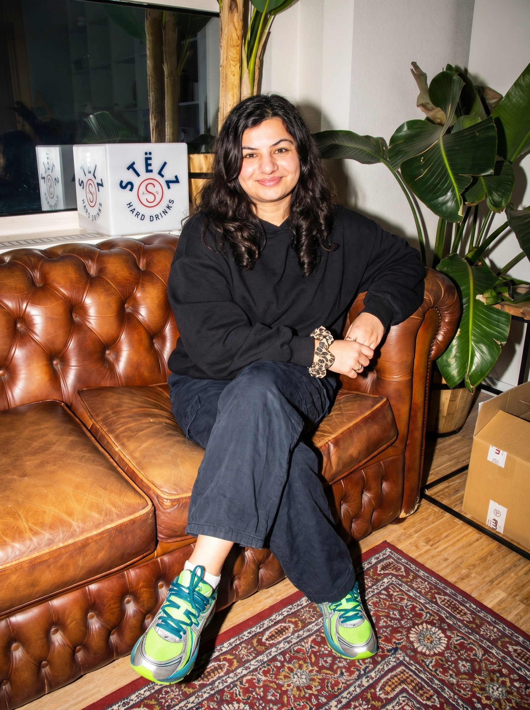 Vrouw in zwarte hoodie en broek met neongroene Asics sneakers op bruine leren chesterfield bank, met Stëlz Hard Drinks branding op de achtergrond