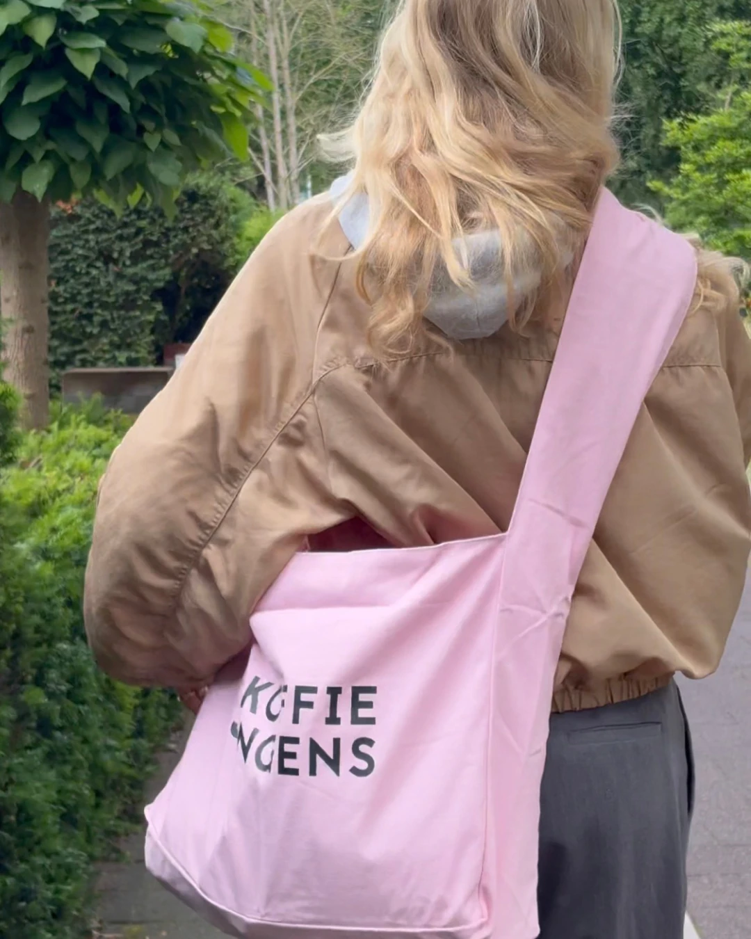 Vrouw draagt de roze tote tas van De Koffiejongens over haar schouder tijdens een wandeling in het park.