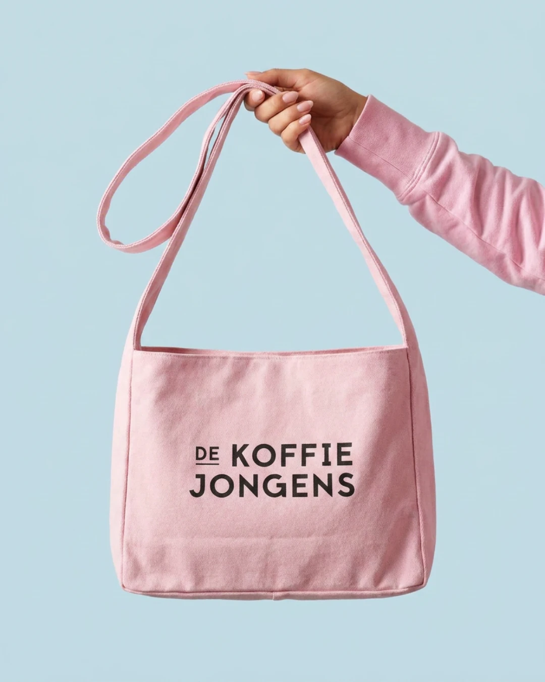 Hand houdt de roze tote tas van De Koffiejongens vast aan het lange hengsel, tegen een blauwe studioachtergrond.