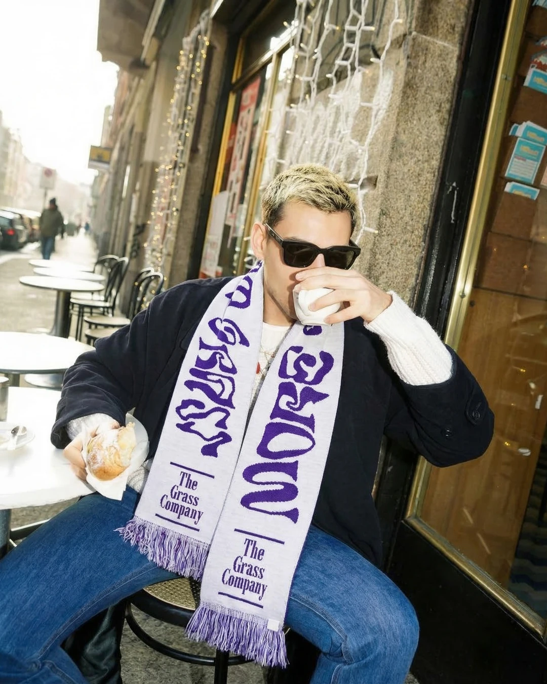 Man draagt de sjaal van The Grass Company op een terras, met een koffie in de hand en een croissant op tafel.