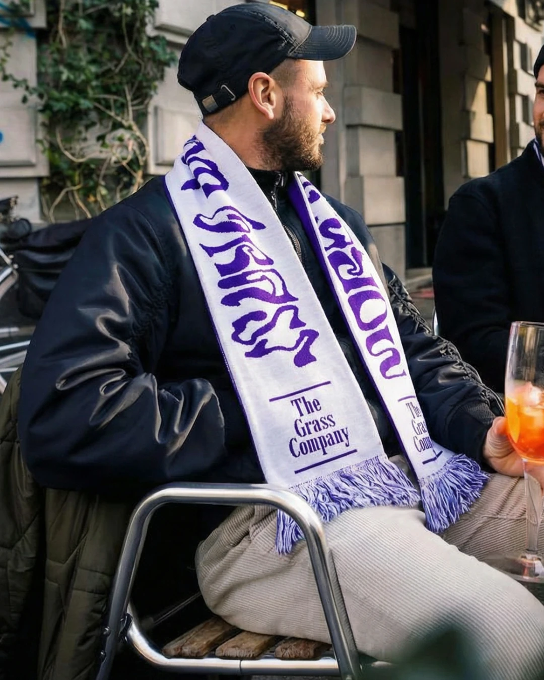 Man met pet draagt de sjaal van The Grass Company op een terras, met een drankje in de hand naast een vriend.